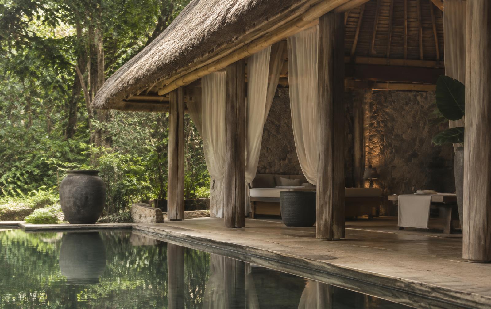 An open-air wellness pavilion with a thatched roof and linen drapes beside a still pool in the jungle